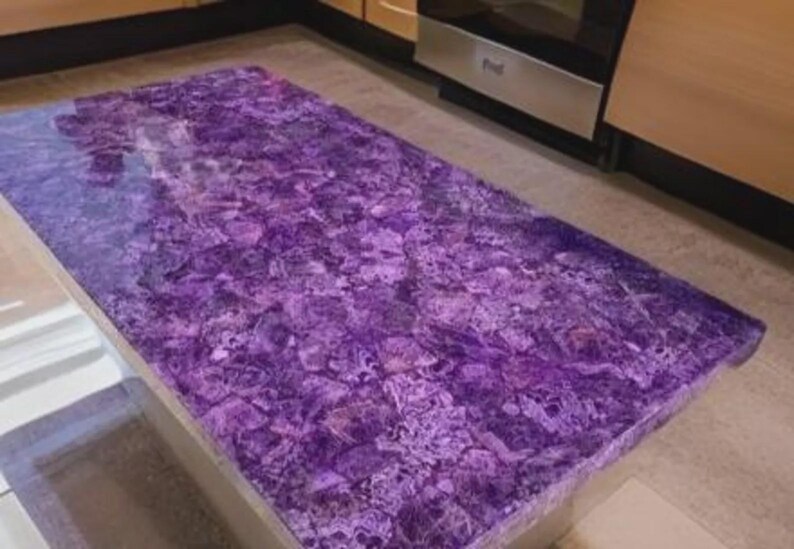 Puede incluir: Una encimera rectangular de amatista con una superficie pulida. La encimera es de color p&uacute;rpura oscuro con vetas p&uacute;rpuras y blancas m&aacute;s claras. La encimera est&aacute; colocada en una cocina con gabinetes de color claro y electrodom&eacute;sticos de acero inoxidable.