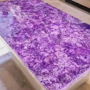 Puede incluir: Una mesa de centro rectangular con una vibrante superficie p&uacute;rpura, similar a un mosaico. La parte superior de la mesa es una pieza s&oacute;lida de amatista, que muestra un patr&oacute;n natural y texturizado. La mesa tiene una base de tono neutro.
