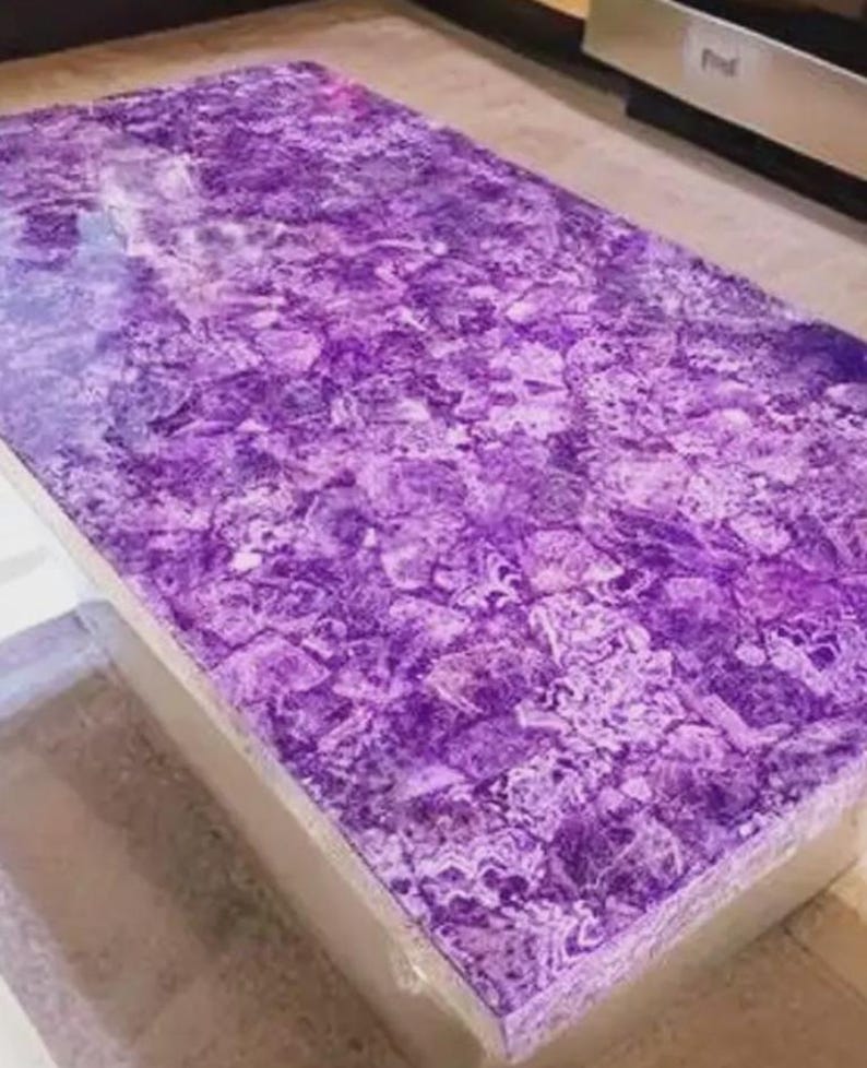 Puede incluir: Una mesa de centro rectangular con una vibrante superficie p&uacute;rpura, similar a un mosaico. La parte superior de la mesa es una pieza s&oacute;lida de amatista, que muestra un patr&oacute;n natural y texturizado. La mesa tiene una base de tono neutro.