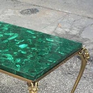 以下が含まれることがあります： マラカイトグリーンの天板と金色の金属製の脚を持つ長方形のコーヒーテーブル。テーブルの天板には、濃い緑と薄い緑の渦巻き模様があります。脚は装飾的で華やかなデザインです。