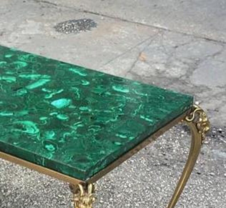 以下が含まれることがあります： マラカイトグリーンの天板と金色の金属製の脚を持つ長方形のコーヒーテーブル。テーブルの天板には、濃い緑と薄い緑の渦巻き模様があります。脚は装飾的で華やかなデザインです。