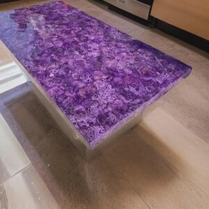 Puede incluir: Una mesa de centro rectangular con una parte superior morada vibrante y estampada. La superficie de la mesa tiene una apariencia de mosaico, con diferentes tonos de morado y blanco. La base parece ser un material transparente y s&oacute;lido.