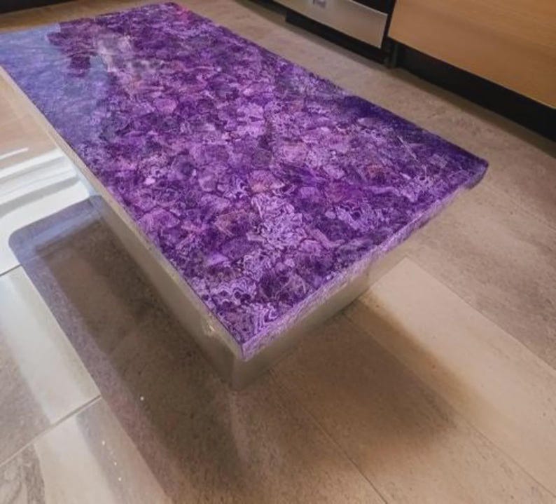 Puede incluir: Una mesa de centro rectangular con una parte superior morada vibrante y estampada. La superficie de la mesa tiene una apariencia de mosaico, con diferentes tonos de morado y blanco. La base parece ser un material transparente y s&oacute;lido.