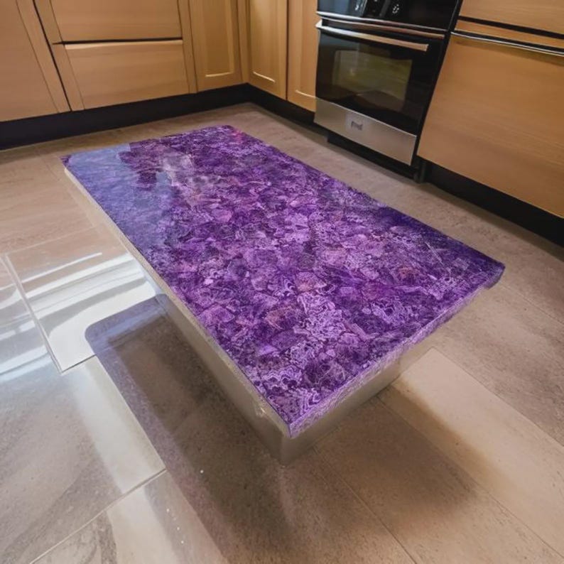 Puede incluir: Isla de cocina rectangular con encimera morada vibrante y texturizada. La encimera tiene un dise&ntilde;o &uacute;nico y estampado. La isla est&aacute; soportada por una base rectangular transparente. La cocina cuenta con gabinetes de madera clara y un horno de acero inoxidable.