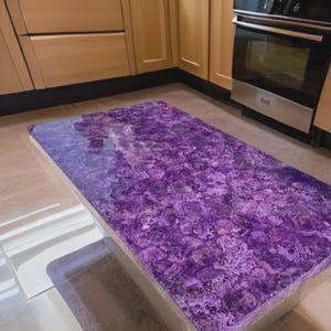 Puede incluir: Una encimera de cocina rectangular con una vibrante superficie p&uacute;rpura texturizada. La encimera se encuentra sobre un fondo de armarios de madera de color claro y un horno de acero inoxidable.