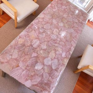 May include: A rectangular coffee table with a pink, mosaic-like surface. The table's top is made of irregularly shaped, light pink stones. The table has short, dark legs and is set on a neutral-colored rug. Two chairs with light-colored seats and wooden frames are visible.