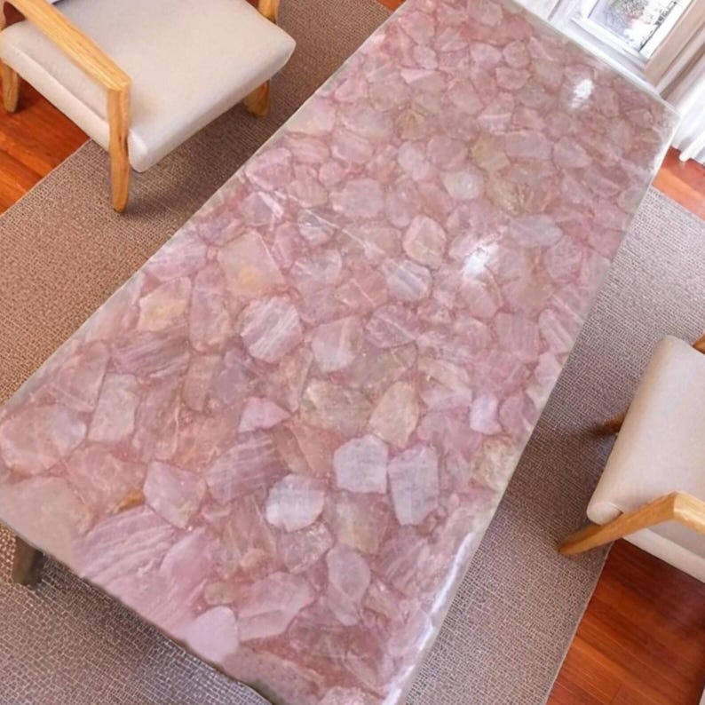 May include: A rectangular coffee table with a pink, mosaic-like surface. The table's top is made of irregularly shaped, light pink stones. The table has short, dark legs and is set on a neutral-colored rug. Two chairs with light-colored seats and wooden frames are visible.