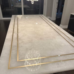 May include: A rectangular white marble table with gold trim. The table has a polished surface and a gold border that frames the edges. A crystal chandelier hangs above the table.