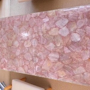 May include: A rectangular table top made of rose quartz. The surface is a mosaic of irregular, light pink and translucent quartz pieces. The table top is a light pink color and has a glossy finish.