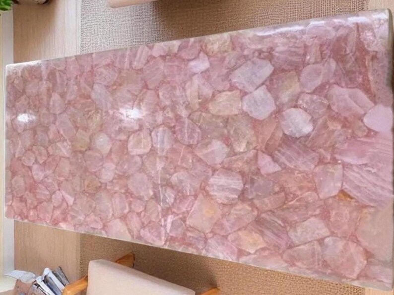 May include: A rectangular table top made of rose quartz. The surface is a mosaic of irregular, light pink and translucent quartz pieces. The table top is a light pink color and has a glossy finish.