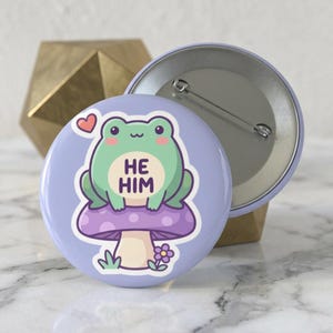 Peut inclure: Un badge rond avec un fond violet clair représentant une grenouille verte assise sur un champignon violet à pois blancs. La grenouille a un petit cœur au-dessus de la tête et le texte HE HIM imprimé sur son torse. Le dos montre une épingle en métal.