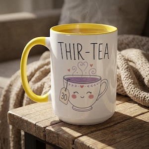 Könnte beinhalten: Ein weißer Keramikbecher mit leuchtend gelbem Henkel und gelbem Innenrand. Auf der Vorderseite steht der schwarze Schriftzug THIR-TEA über einer Grafik einer lächelnden Teetasse mit einem Teebeutel mit der Aufschrift 30, umgeben von violetten Dampfwirbeln.