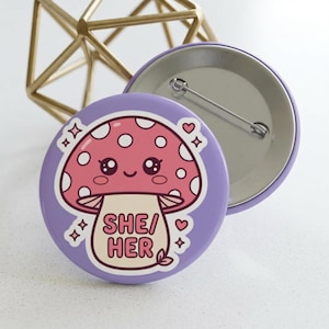 Puede incluir: Una chapa redonda con fondo morado claro que presenta un personaje de seta rosa con puntos blancos, una cara sonriente y el texto SHE/HER en letras rosas. El diseño incluye pequeños corazones rosas y detalles de estrellas blancas.