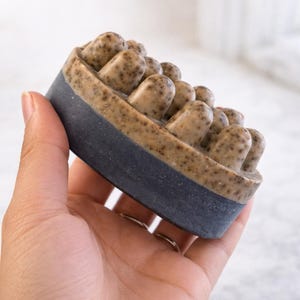 May include: A dark blue and tan soap bar with a textured surface. The top of the soap has multiple rounded protrusions. The soap is held in a hand.