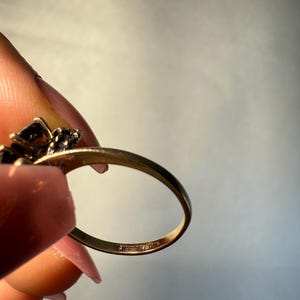 以下が含まれることがあります： 暗い宝石のクラスターが付いたゴールドリング。 リングバンドは細く、「SIFFARI 14」の刻印があります。 リングは明るい背景に対して保持されています。