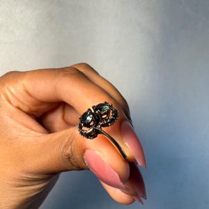 以下が含まれることがあります： 2つの濃い緑色の宝石をあしらったゴールドトーンのリング。リングのデザインには、石の周りの装飾的なディテールが含まれています。リングは、長いピンク色の爪を持つ人によって保持されています。