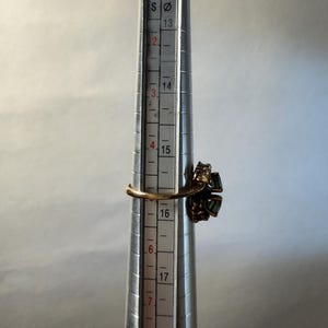 以下が含まれることがあります： 緑色の宝石が付いたゴールドトーンのリングがリングサイザーに置かれています。リングのサイズは約16号です。リングサイザーはシルバーで、黒いマーキングがあります。