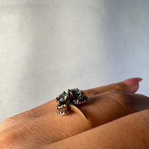 以下が含まれることがあります： 2つの楕円形の濃い緑色の宝石をあしらったゴールドリング。リングは、石の周りに複雑なディテールを持つユニークなデザインです。リングは指に表示され、そのエレガントで洗練されたスタイルを強調しています。