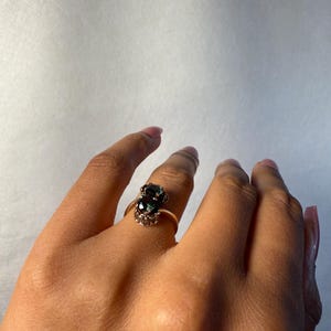 以下が含まれることがあります： 2つの楕円形の濃い緑色の宝石をあしらったゴールドリング。リングは、メインの宝石を囲む小さなクリアストーンを備えた、繊細で装飾的なセッティングです。マニキュアされた爪のある手にリングが表示されています。