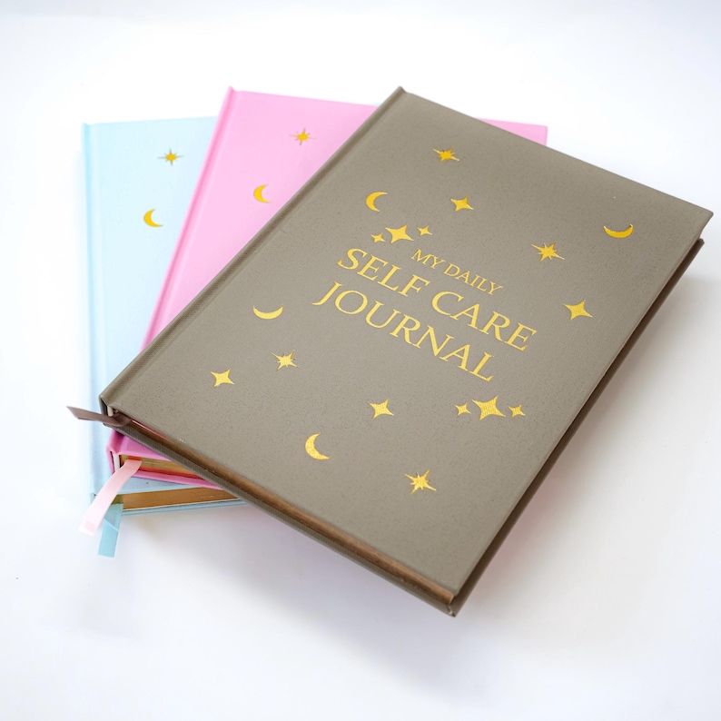 May include: Three journals in pastel colors: light blue, pink, and gray. The gray journal is on top, with gold text that reads "MY DAILY SELF CARE JOURNAL." Each journal has gold stars and crescent moon designs.
