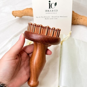May include: A wooden scalp massager with a smooth, contoured handle and a circular head featuring numerous metallic copper-coloured rounded pins. Behind the massager is a white card with the text "ic IRKA & CO GLOW RITUALS Spa & Tea Rituals - Botanical Wellness Made with Love."