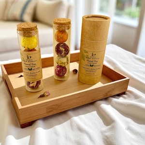 May include: A wooden tray holds two glass tubes with cork stoppers, each containing floral tea balls. A third, cylindrical container of floral tea sits on the tray. The tea balls are various colors, including yellow, orange, and red. The text on the label reads "Floral Tea Calm & Relax 100% Natural".