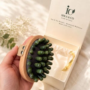 Op de afbeelding: Een handmatige massageborstel met een lichte houten basis, een beige stoffen band en talloze donkergroene, jade-achtige cilindrische noppen. De borstel wordt gepresenteerd naast een open crèmekleurige doos met het label IRKA & CO GLOW RITUALS.
