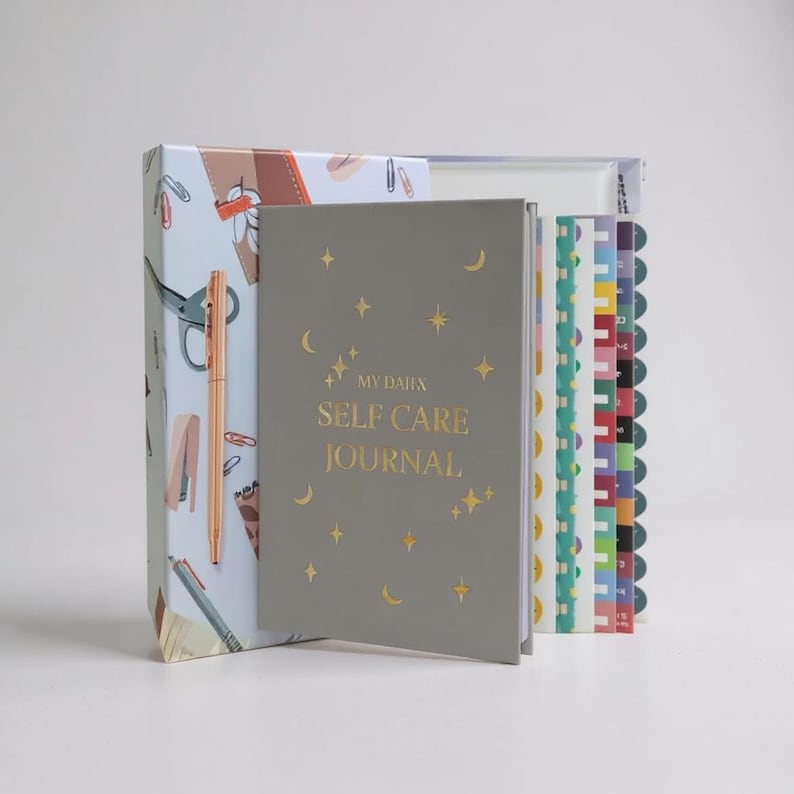 May include: A gray self-care journal with gold stars and moons, the text "MY DAILY SELF CARE JOURNAL" is printed on the cover. A pen and a patterned box are visible in the background, along with a set of colorful notebooks.