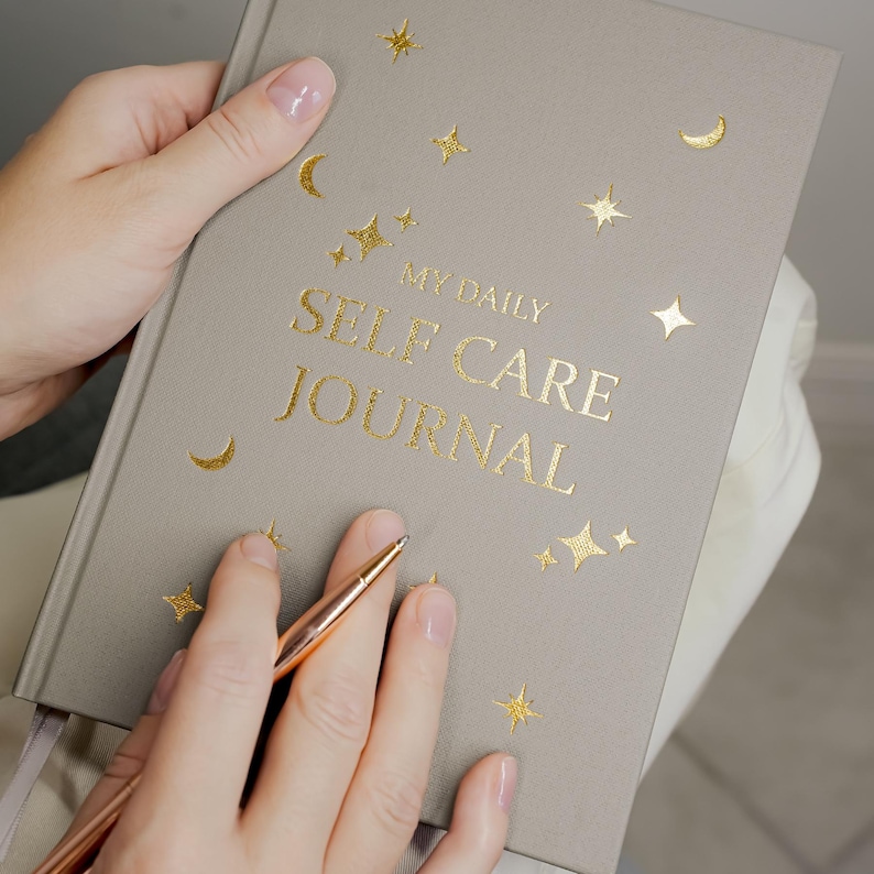 May include: A gray journal with gold foil lettering that reads "MY DAILY SELF CARE JOURNAL." The journal is decorated with gold stars and crescent moons. A rose gold pen rests on the journal.