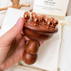 May include: A handheld wooden massage tool with a rounded handle and a flat circular head featuring multiple small, polished copper-colored metal spheres embedded on the surface. The tool is presented in a box with text reading IRKA & CO.