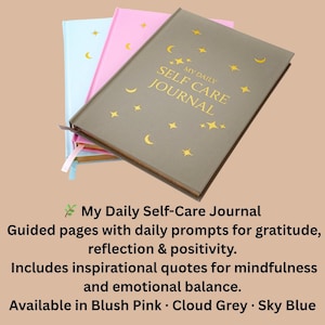 May include: Three journals in cloud grey, blush pink, and sky blue. The cloud grey journal is in the foreground, with gold text that reads "My Daily Self Care Journal". The journal has gold stars and crescent moon designs.