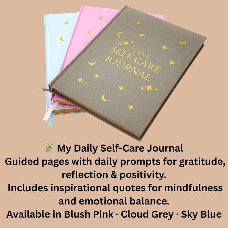 May include: Three journals in cloud grey, blush pink, and sky blue. The cloud grey journal is in the foreground, with gold text that reads "My Daily Self Care Journal". The journal has gold stars and crescent moon designs.