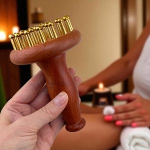 Op de afbeelding: Een houten massagegereedschap met een afgeronde kop en een handvat. De kop heeft meerdere goudkleurige, cilindrische massagepunten. Het gereedschap wordt in een hand gehouden, met een vage achtergrond van een spa-omgeving.