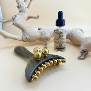 Könnte beinhalten: Ein Massage-Tool aus Holz mit goldfarbenen Metallkugeln und einer Ölflasche mit einer Pipette. Auf der Ölflasche steht "Glow Ritual Multi-Use Oil Neroli Infused". Der Hintergrund zeigt einen hellfarbigen, strukturierten Ast.