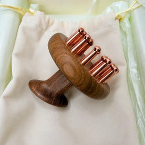 Könnte beinhalten: Ein manuelles Massagegerät mit einem dunkelbraunen Holzgriff und einer kreisförmigen Basis, von deren Oberseite mehrere glatte, kupferfarbene Metallstifte abstehen. Das Gerät liegt vor einem schlichten, hellen Stoffbeutel.