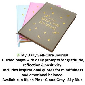 May include: Three journals in blush pink, cloud grey, and sky blue. The cloud grey journal is on top, with gold text reading "MY DAILY SELF CARE JOURNAL" and gold star and crescent moon designs. The text below says "My Daily Self-Care Journal".