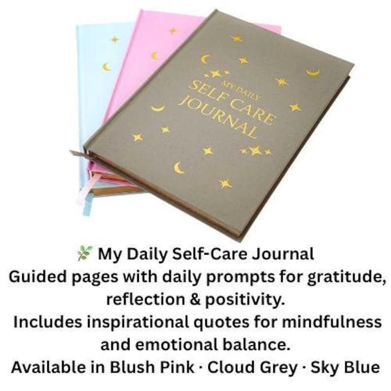 May include: Three journals in blush pink, cloud grey, and sky blue. The cloud grey journal is on top, with gold text reading "MY DAILY SELF CARE JOURNAL" and gold star and crescent moon designs. The text below says "My Daily Self-Care Journal".