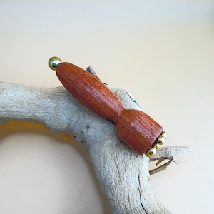 Puede incluir: Un tapón de botella de madera hecho a mano con un acabado marrón rojizo. El tapón tiene un cuerpo redondeado y está adornado con pequeñas esferas metálicas doradas en la base y una sola esfera en la parte superior. Descansa sobre un trozo de madera flotante.