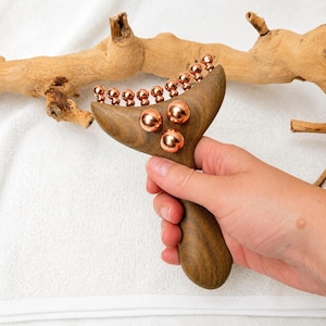 Op de afbeelding: Een handmatig houten massagehulpmiddel met een gebogen, Y-vormige kop, voorzien van een rij kleine koperkleurige metalen bolletjes langs de bovenrand en drie grotere koperkleurige metalen bolletjes in het midden van het donkerbruine hout.