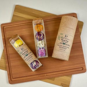 May include: Three glass tubes with cork stoppers, each containing colorful floral tea balls. One tube has a yellow tea ball, another has orange and purple tea balls, and the third has pink and white tea balls. A brown paper tube and a wooden cutting board are also in the image.