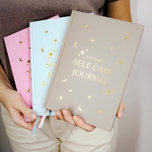 May include: Three journals in pink, light blue, and taupe are held in front of a person. The taupe journal has the text "MY DAILY SELF CARE JOURNAL" in gold lettering, along with gold stars and crescent moon designs. The other journals have the words "SELF CARE" in gold.