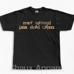 Può includere: T-shirt nero con la scritta "never stressed im dead calm" in un carattere dorato con stampa leopardata. La maglietta è del marchio Gildan. Il testo è centrato sulla parte anteriore della maglietta.