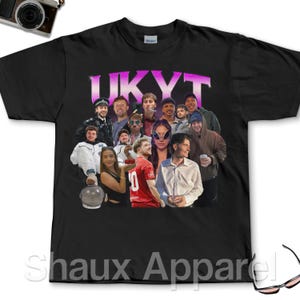 Puede incluir: Camiseta negra con un grupo de personas y el texto "UKYT" en degradado rosa y morado. La camiseta tiene cuello redondo y mangas cortas. La marca "Gildan" es visible en el cuello.