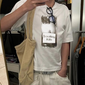 May include: White t-shirt with a graphic of a phone case that says "Scrolling Kills." The shirt is paired with white pants, a beige corduroy tote bag, and black eyeglasses.