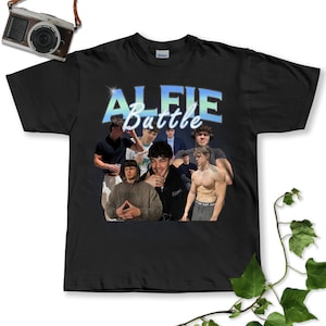 May include: Black t-shirt featuring a collage of images and the text "ALFIE Battle" in a blue and white design. The shirt has a crew neck and short sleeves. The images are of a young man in various poses.