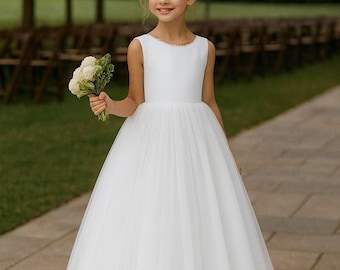 Op de afbeelding: Een jong meisje in een witte mouwloze jurk met een tule rok, met een boeket witte bloemen. De jurk heeft een met kralen bezette halslijn. Het meisje glimlacht, met haar haar in een updo. De achtergrond is een wazige buitenomgeving.