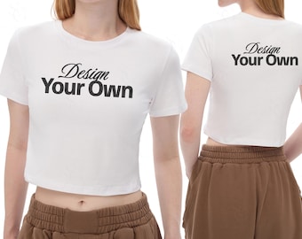 Może przedstawiać: Dwa białe topy typu crop z czarnym napisem "Design Your Own" z przodu i z tyłu. Koszulki mają krótkie rękawy i okrągły dekolt. Modelka ma na sobie brązowe szorty.