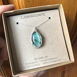 Peut inclure: Un collier en argent avec un pendentif en labradorite. Le pendentif est enveloppé de fil d'argent et la pierre est de couleur bleu-vert avec des reflets irisés. La boîte dans laquelle se trouve le collier porte les mots "Labradorite" et "Pierre de force - Persévérance, Coïncidence - Synchronisation" imprimés dessus.