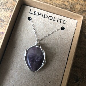 May include: A silver chain necklace with a purple lepidolite stone pendant wrapped in silver wire. The necklace is displayed in a brown box with the word "LEPIDOLITE" printed on the lid.
