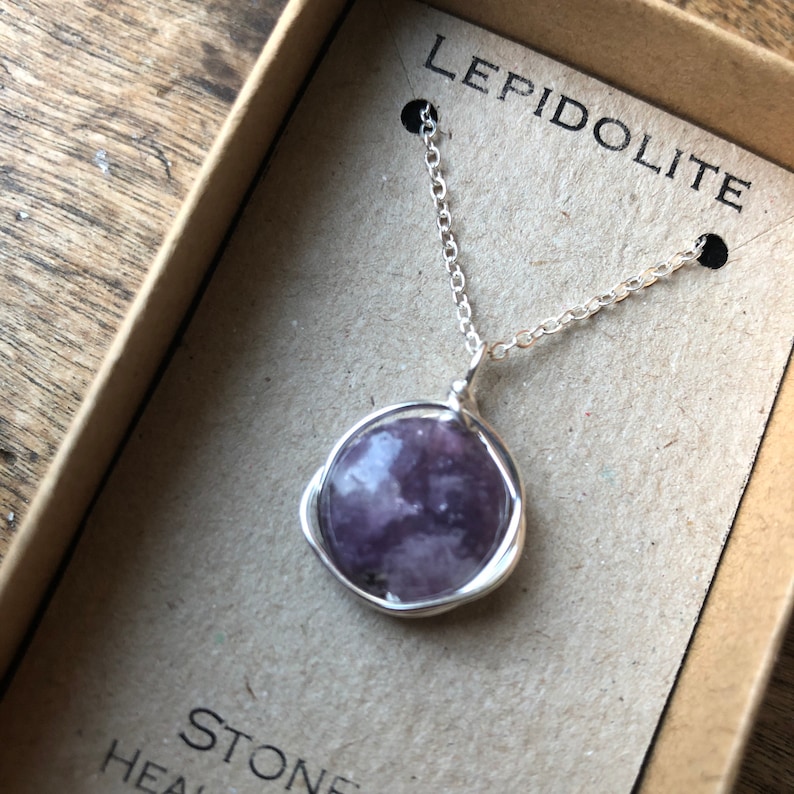 Pu&ograve; includere: Una collana con catena d'argento con un ciondolo in pietra di lepidolita viola. La pietra &egrave; avvolta in filo d'argento e ha un aspetto naturale e nuvoloso. La collana &egrave; esposta in una scatola con la scritta "LEPIDOLITE" stampata su di essa.
